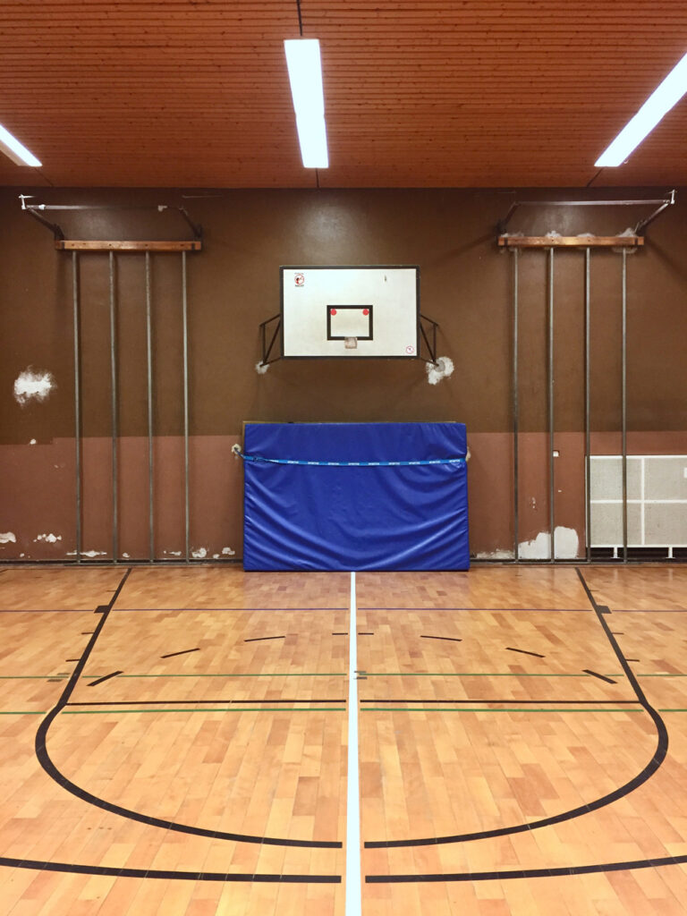 Eine Turnhalle. Auf dem Bild sind eine braune Wand mit zwei Kletterstangen aus Metall mit jeweils vier Stangen. In der Mitte hängt ein Basketballkorb, darunter steht eine große blaue Matte. Der Boden ist Parkett.
In die Holzdecke sind rechteckige Lampen eingelassen.