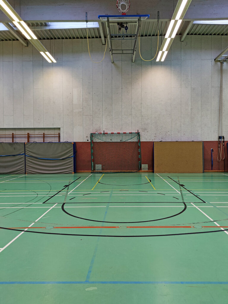 Eine Turnhalle mit türkisfarbenem Linoleumboden. Darauf sind mehrere sich kreuzende schwarze, rote, blaue, gelbe und weiße Linien und Kreise aufgemalt.
Die Wand besteht aus hellgrauem Beton und einem unteren Teil aus rostrotem Filz. Dort stehen ein Handballtor, mehrere weiche Matten, Sprossenwände aus Holz sowie Klettertaue.
An der Decke hängt ein eingeklappter Basketballkorb und die Halle wird von Neonröhren beleuchtet. Sie wirkt insgesamt etwas heruntergekommen.